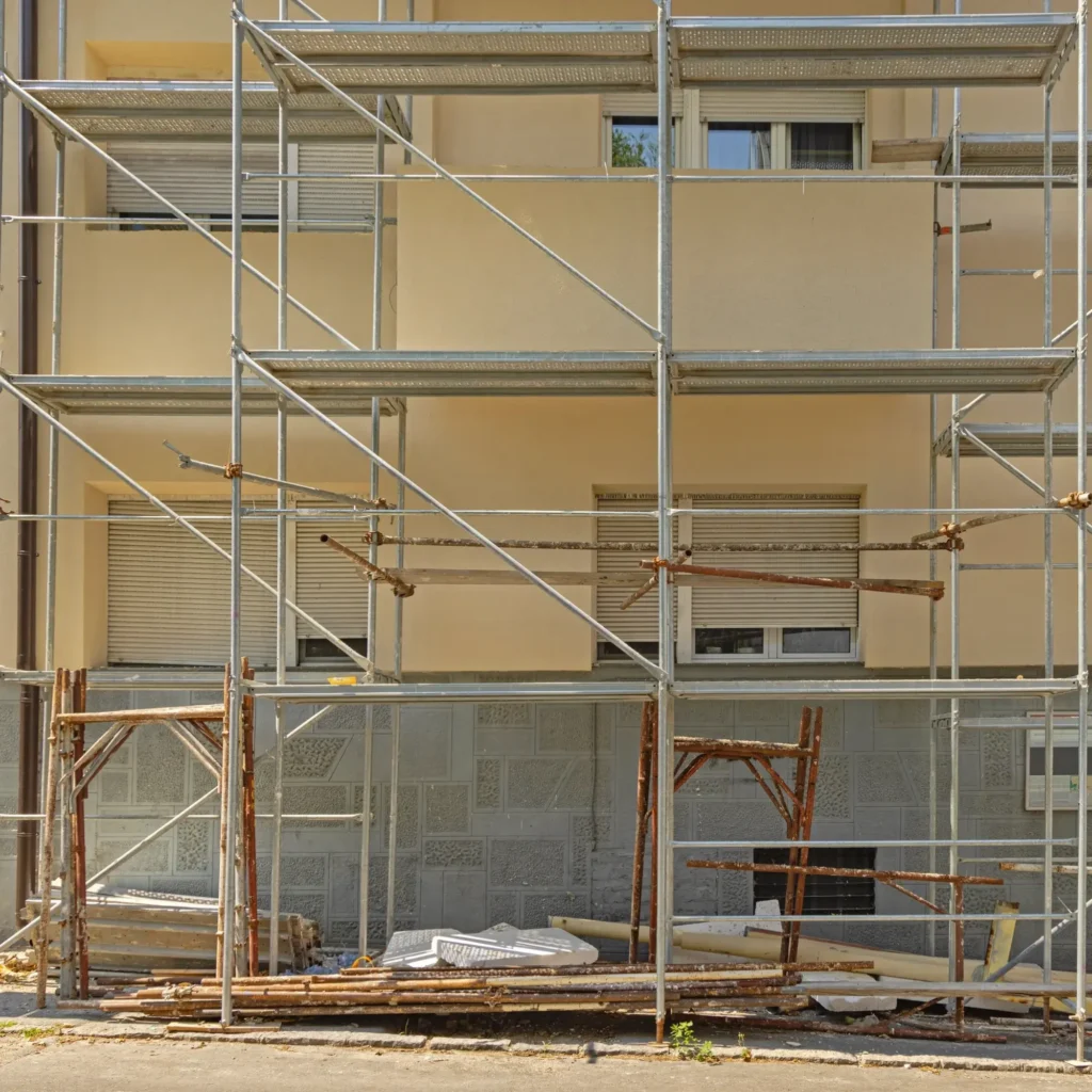 Rénovation de façade : échafaudage et matériaux de construction. Scène de rénovation : grand échafaudage métallique sur une façade beige avec volets roulants gris. Matériaux et débris de construction empilés au sol.