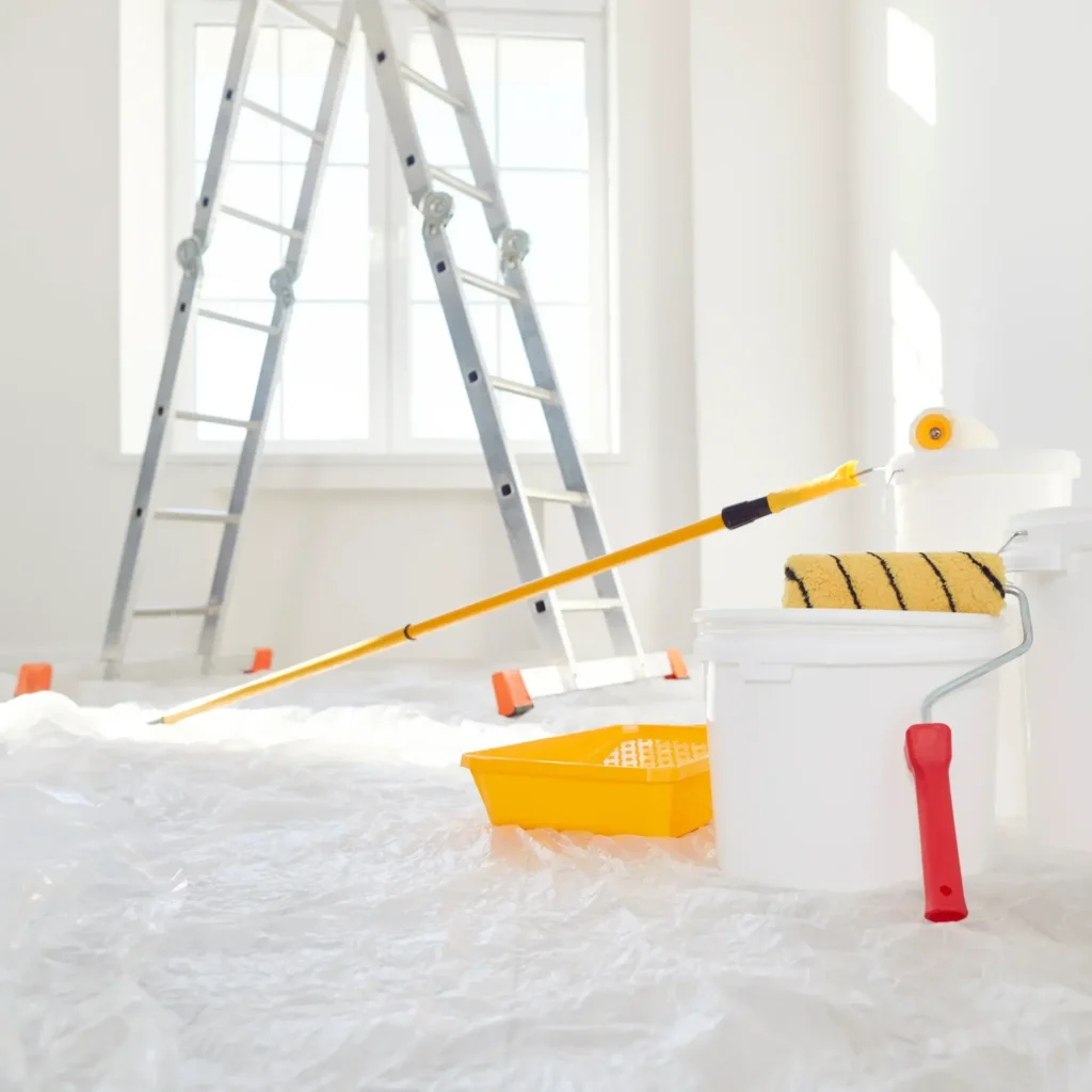 Peinture & Rénovation : Matériel et outils pour vos travaux. Matériel de peinture et rénovation : rouleaux, seaux, perche jaune et bac. Échelle devant une fenêtre, sol protégé par une bâche.
